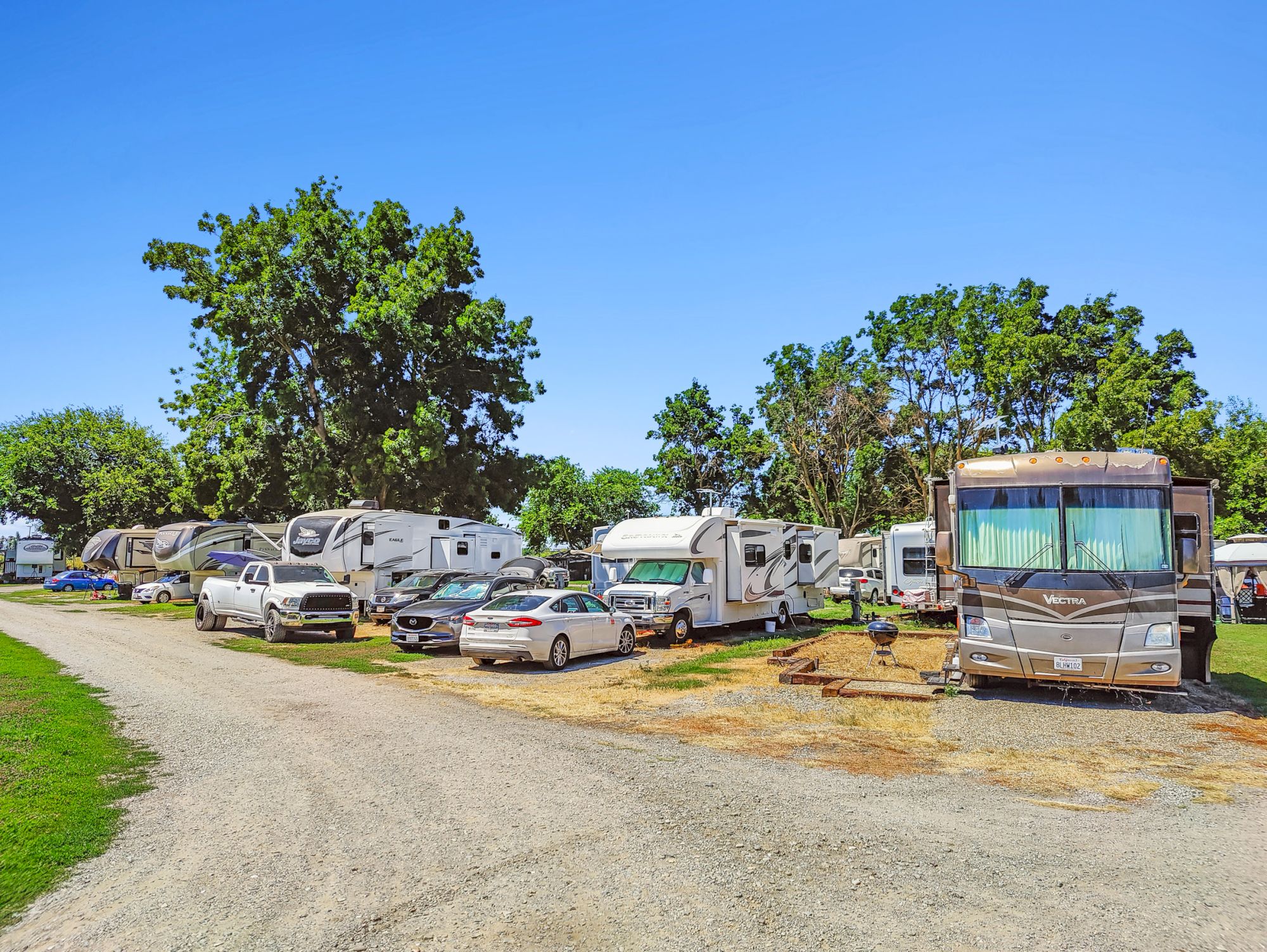 The image shows a campground with parked RVs, a few cars, and trees under a clear blue sky, with a gravel road in the foreground.