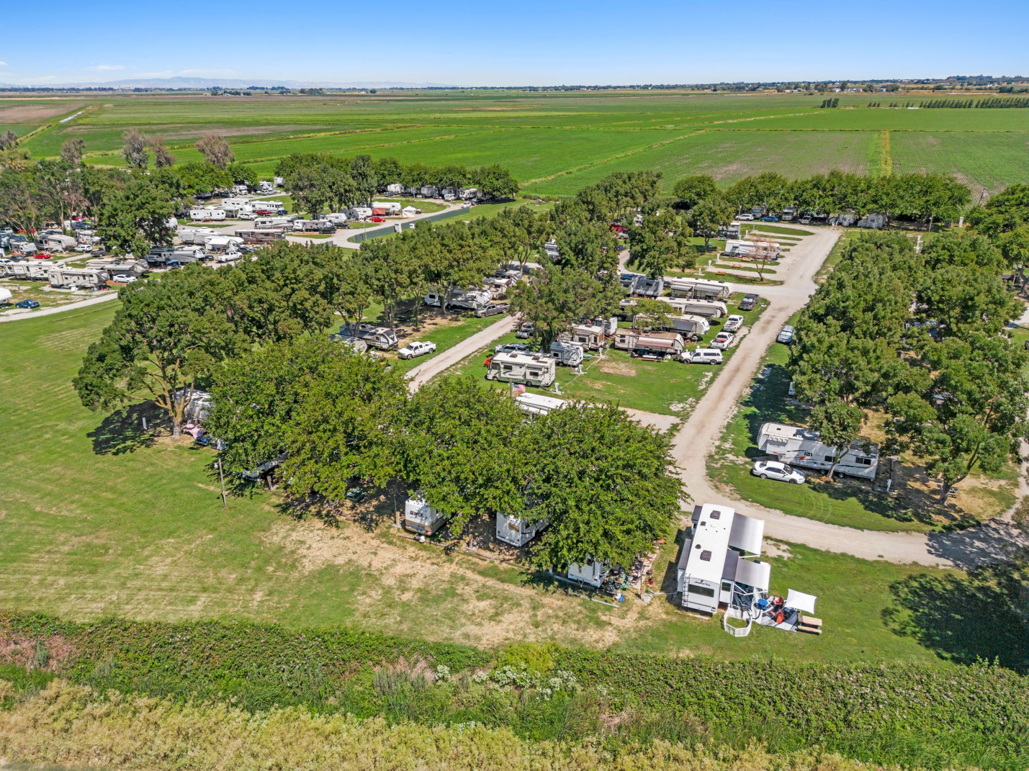 The image shows an aerial view of a campground with RVs and trees arranged along winding roads amidst green fields.