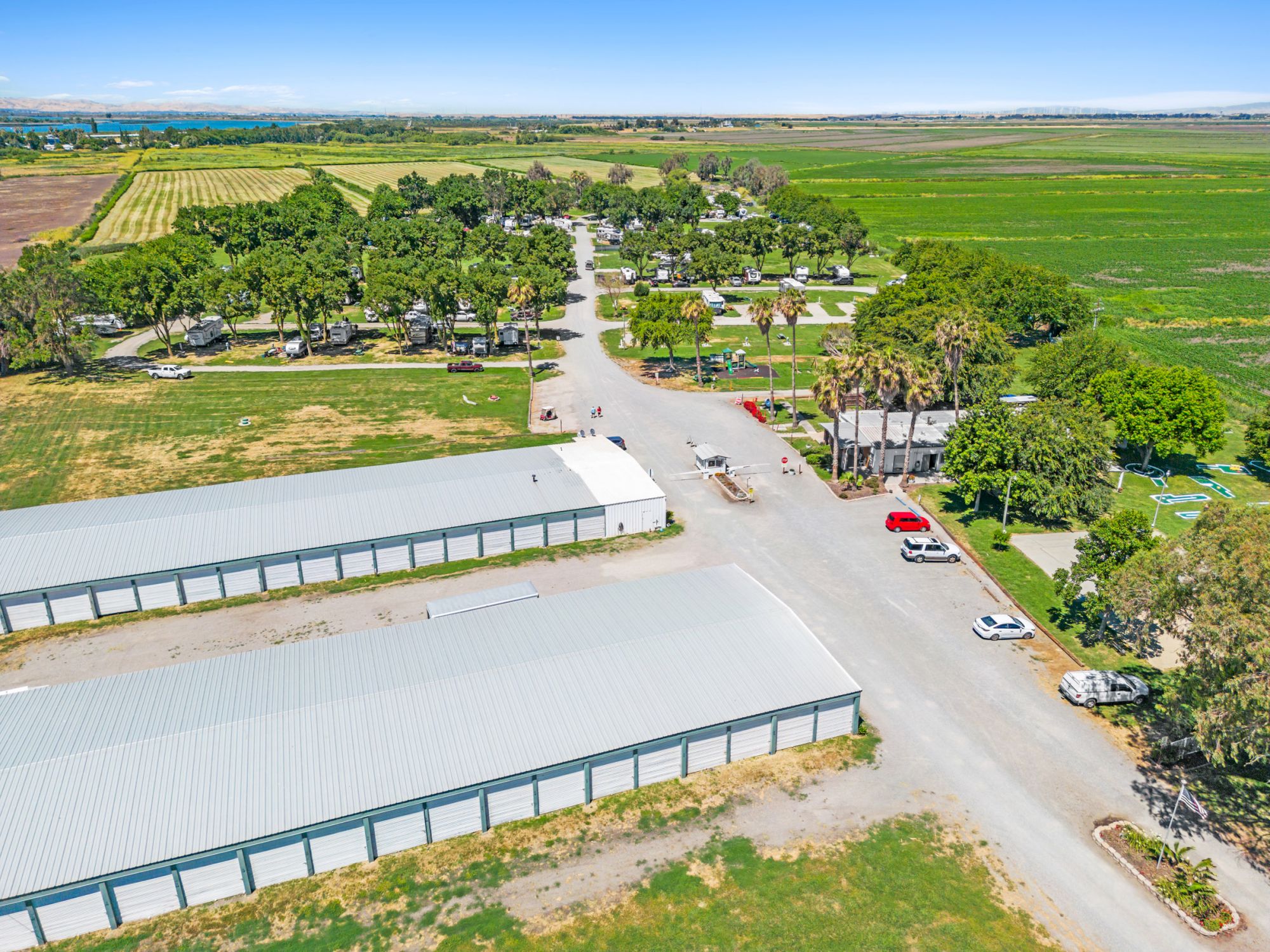 An aerial view shows storage buildings, parked cars, and surrounding greenery in a rural landscape under a clear blue sky.