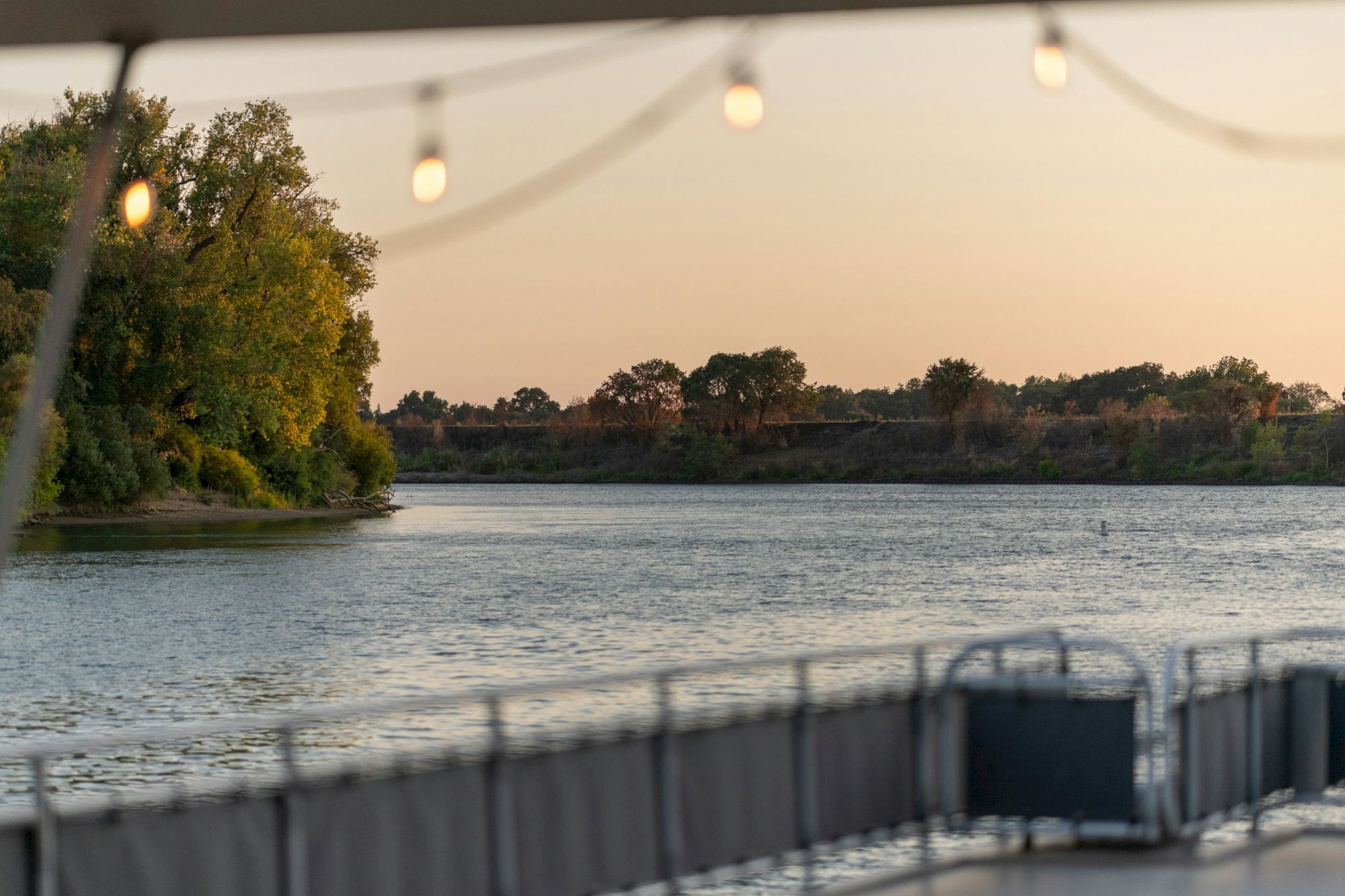 River scene at sunset with trees lining the shore. String lights are visible, suggesting a relaxing outdoor setting on a deck.