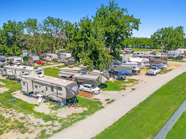 The image shows a campground with multiple RVs parked on grassy plots, surrounded by trees and parked cars along a gravel road.