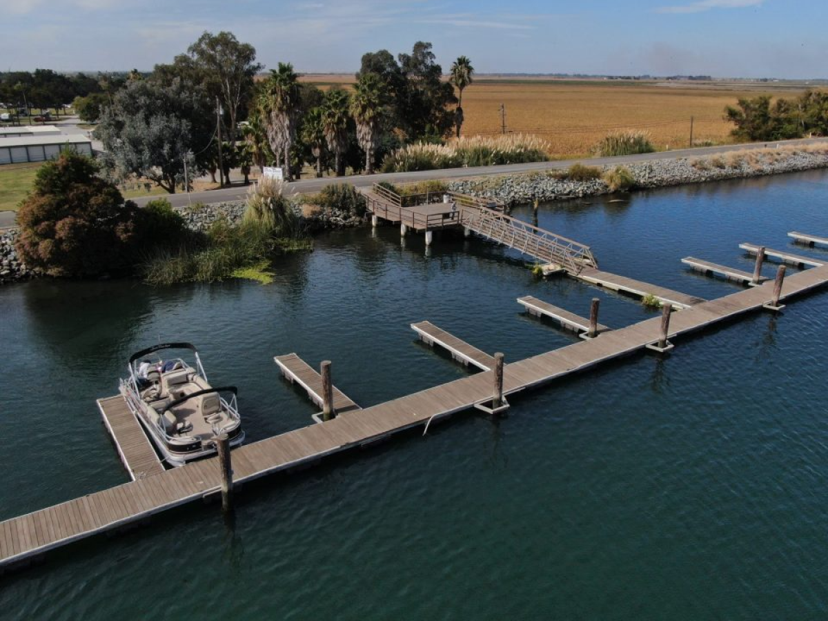 A boat is docked at a wooden pier with multiple slips on a calm body of water, surrounded by trees and open fields in the background.