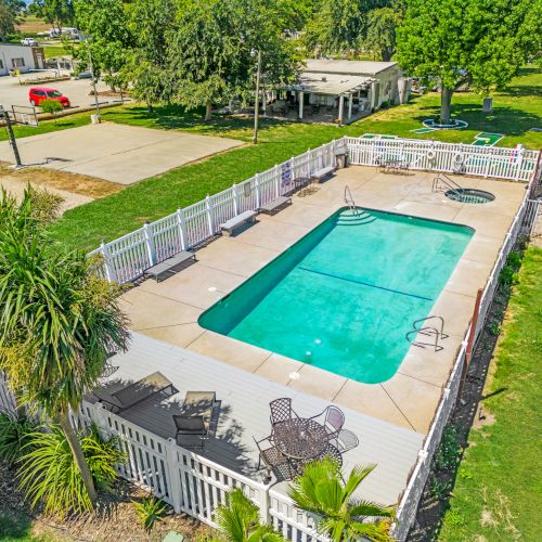 An aerial view of a fenced outdoor swimming pool with deck chairs and surrounding greenery in a spacious yard.