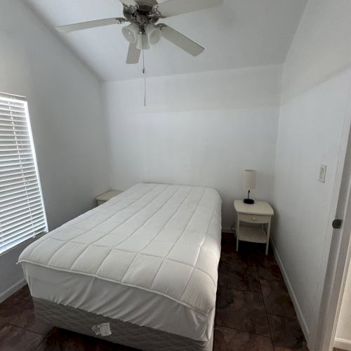 A bedroom with a ceiling fan, a bed with white bedding, two nightstands, a lamp, and a window with blinds.