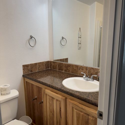 The image shows a bathroom with a sink, wooden cabinets, toilet, and mirror with lights, featuring tiled accents on the countertop.