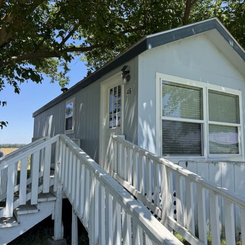 The image shows a small, light blue house with a ramp, windows, and trees surrounding it, under a clear blue sky.