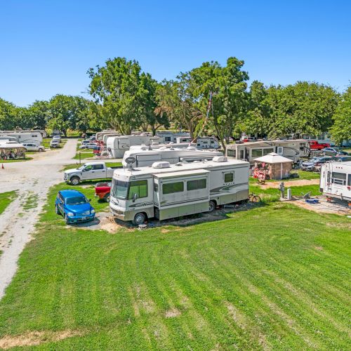 An RV park with several motorhomes and trailers on grassy lots, surrounded by trees and clear skies, creating a serene outdoor setting.