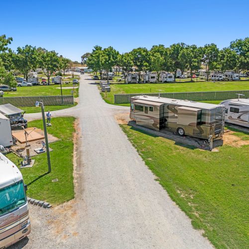 An RV park with several motorhomes, green lawns, and trees under a clear blue sky. A gravel road runs through the center.