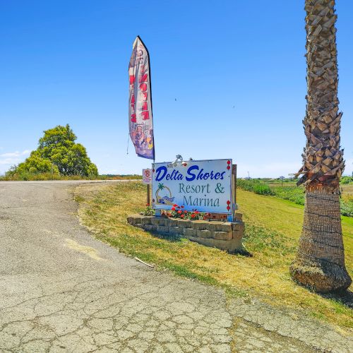 A sign reads "Delta Shores Resort & Marina" on a grassy area beside a road, with a palm tree and a vertical flag nearby.