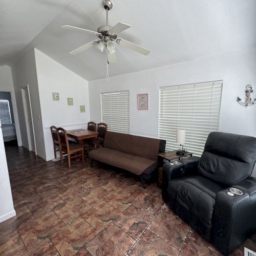 A cozy living room with a ceiling fan, brown sofa, dining table, and black recliner on a tiled floor with two windows and decor.