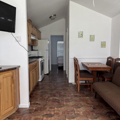 The image shows a small kitchenette and dining area with wood cabinets, a table, and chairs, leading to a bedroom, all in a compact layout.
