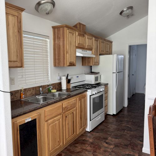 A compact kitchen with wooden cabinets, a gas stove, a refrigerator, and a sink, featuring brown flooring and white walls.