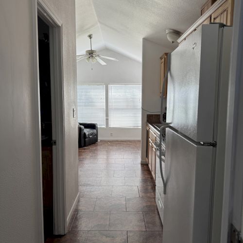 A narrow kitchen hallway with a refrigerator, leading to a bright living area featuring a ceiling fan and large windows at the end.