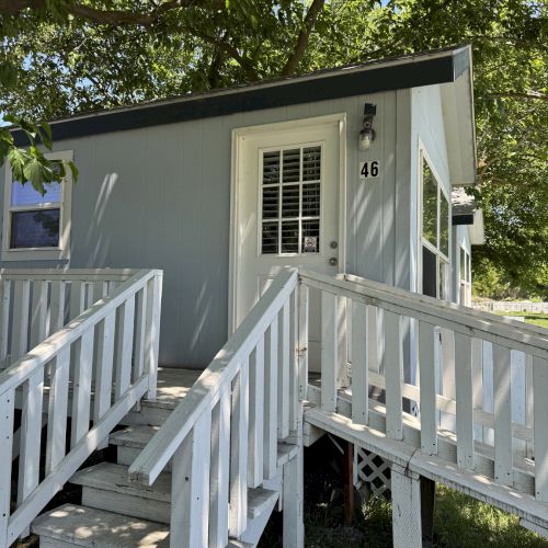 A small light blue cabin with white railings and stairs, surrounded by trees. The door has a light above it and a number 46 beside it.