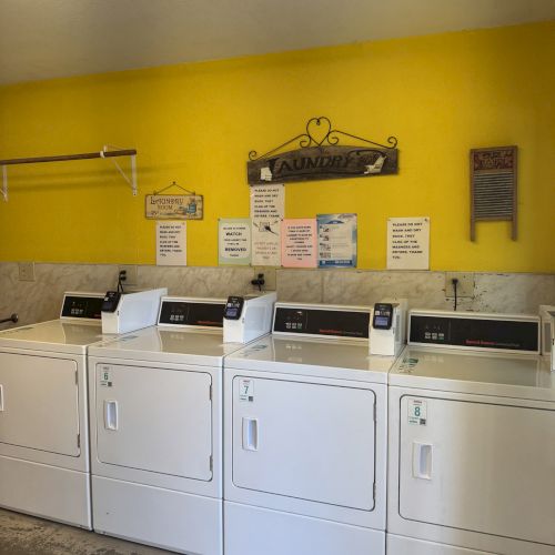 The image shows a laundry room with four white washing machines against a yellow wall, with signs and decor above them.