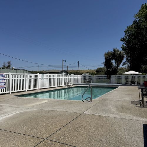 The image shows an outdoor swimming pool surrounded by a white fence, with clear skies and trees in the background.