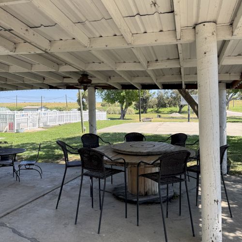 A covered outdoor area with a round table and chairs on a concrete floor, surrounded by greenery and a white fence.