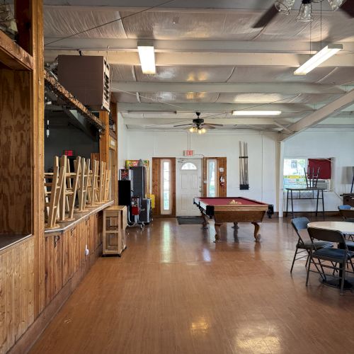 The image shows a room with a pool table, chairs, tables, ceiling fans, and fluorescent lights on a wooden floor, creating a casual setting.