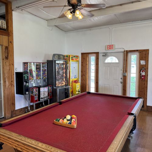 A room with a red pool table, a ceiling fan, arcade machines, and a door.