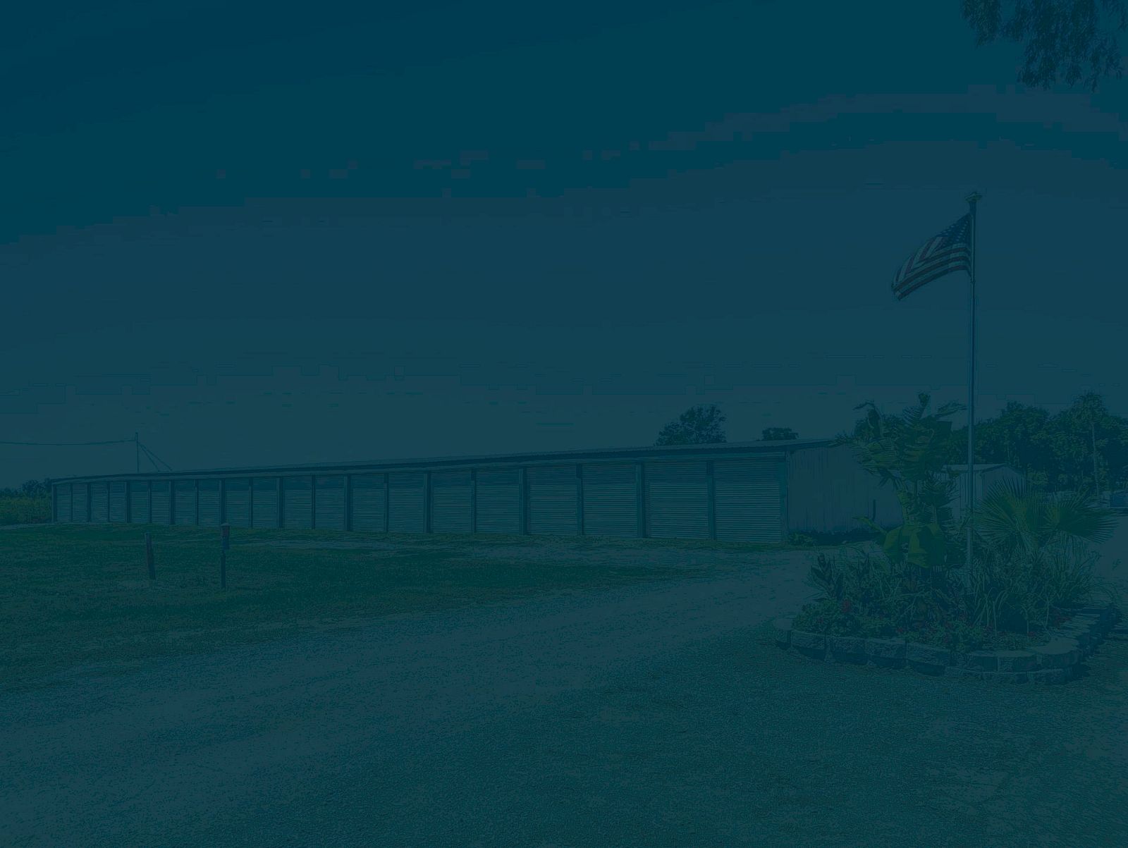 A flag on a pole beside a building with a covered walkway is surrounded by grass and a dirt path.
