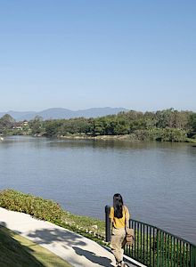 A person stands near a river, looking at the water, with a clear blue sky and mountains in the background.