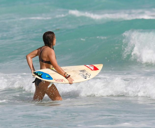 A person carrying a surfboard walks into the ocean, ready to surf the waves, under a clear sky.