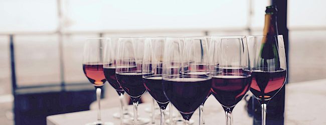 A row of wine glasses filled with red wine sits on a table, with a bottle of wine nearby in a bright setting.