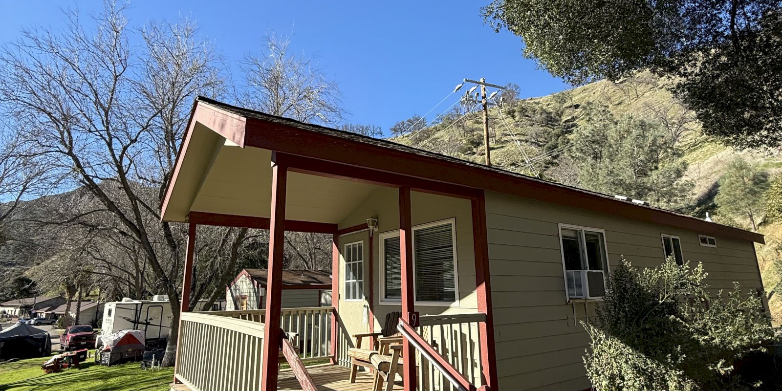 A small, yellow cabin with a red porch is set on a grassy area surrounded by trees and hills under a clear blue sky.