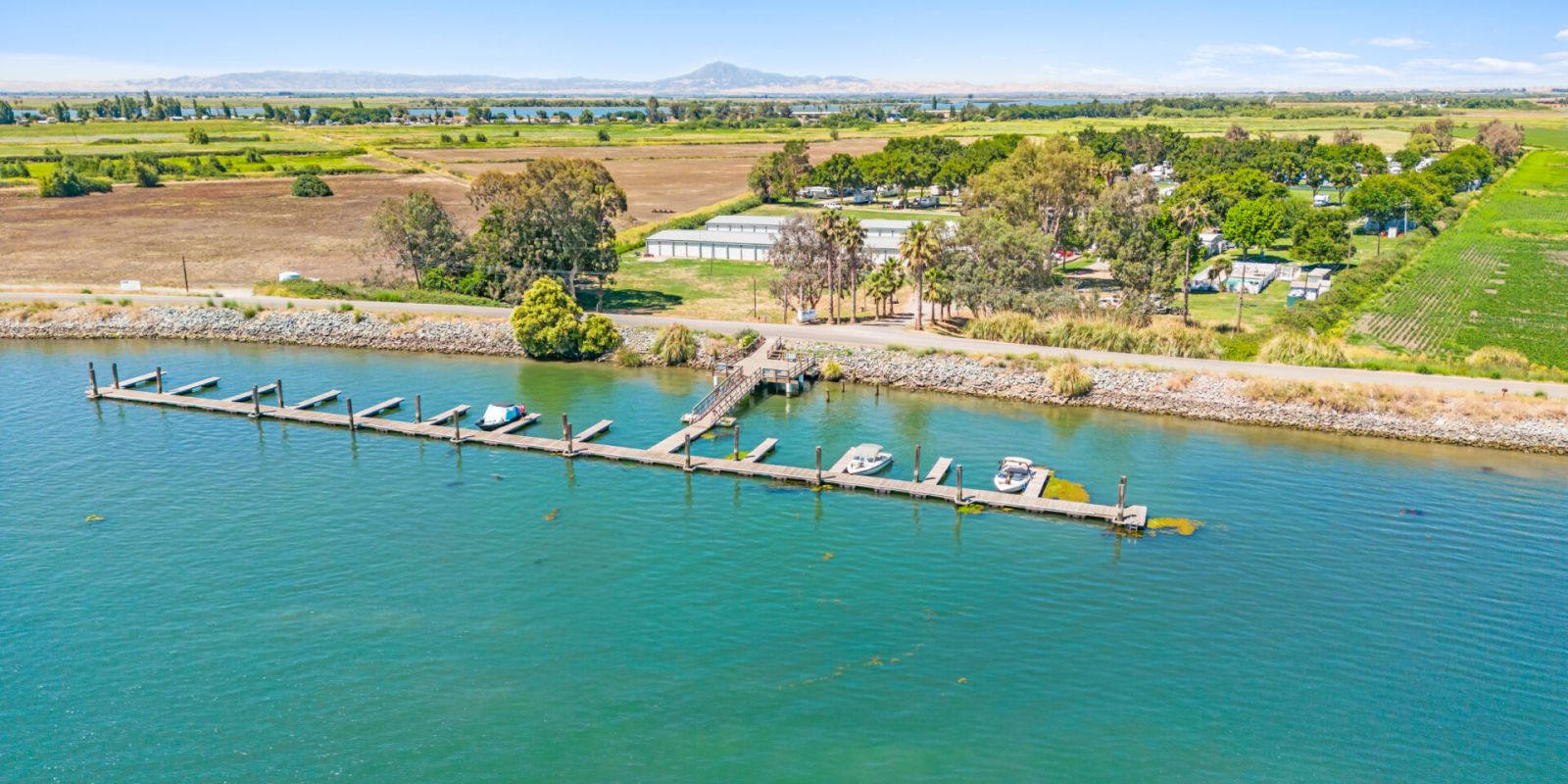 A serene landscape with a long dock extending into a calm body of water, surrounded by lush greenery and fields under a clear blue sky.