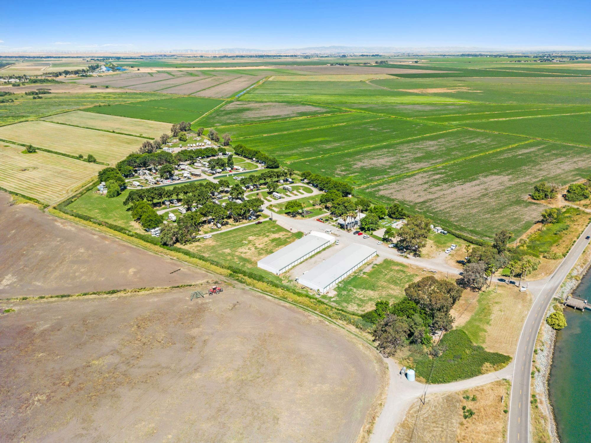 The image shows an aerial view of a rural landscape with green fields, a cluster of trees, buildings, and a nearby road by a body of water.