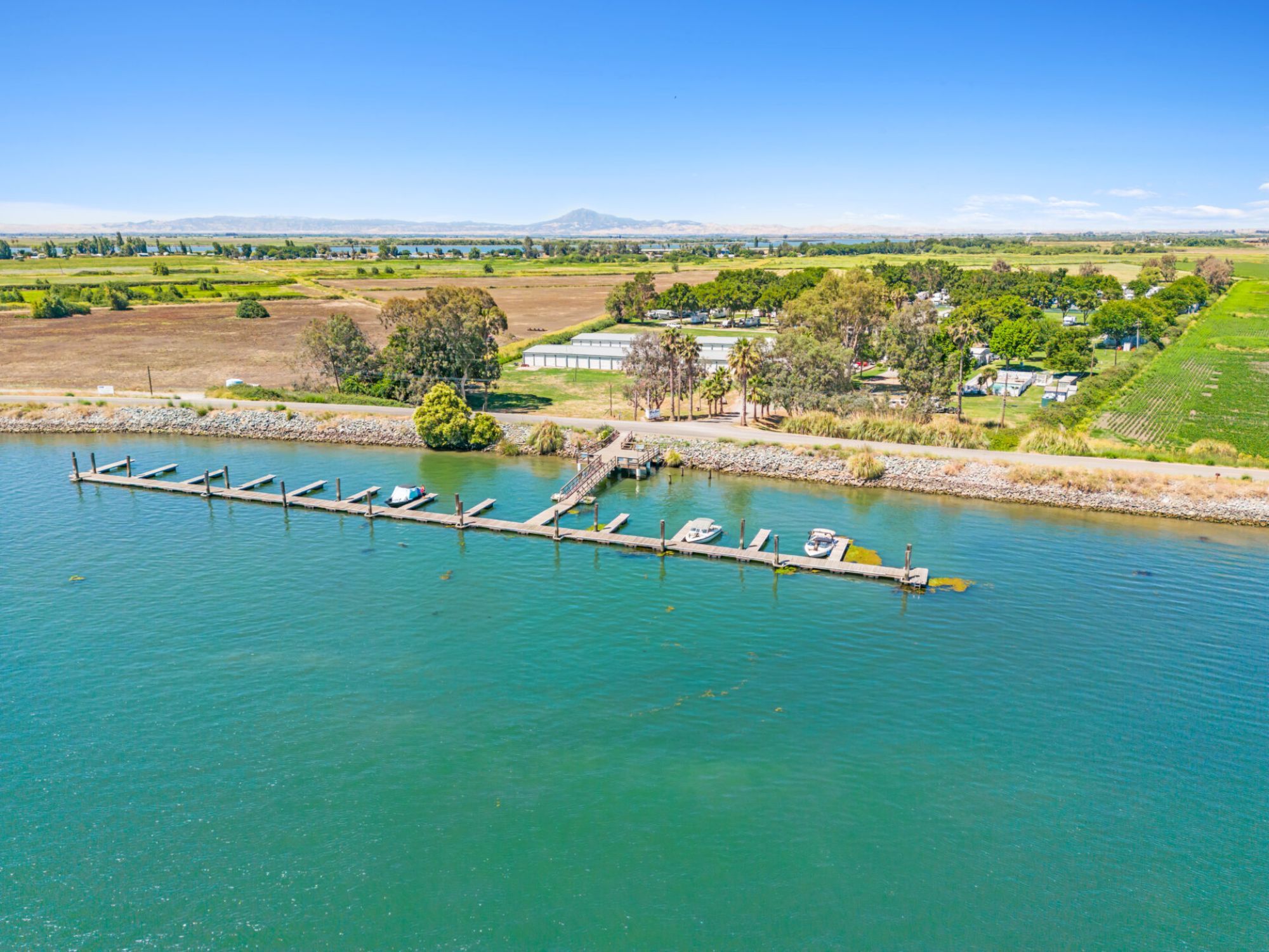Aerial view of a harbor with boats docked, surrounded by greenery and fields, under a clear blue sky.